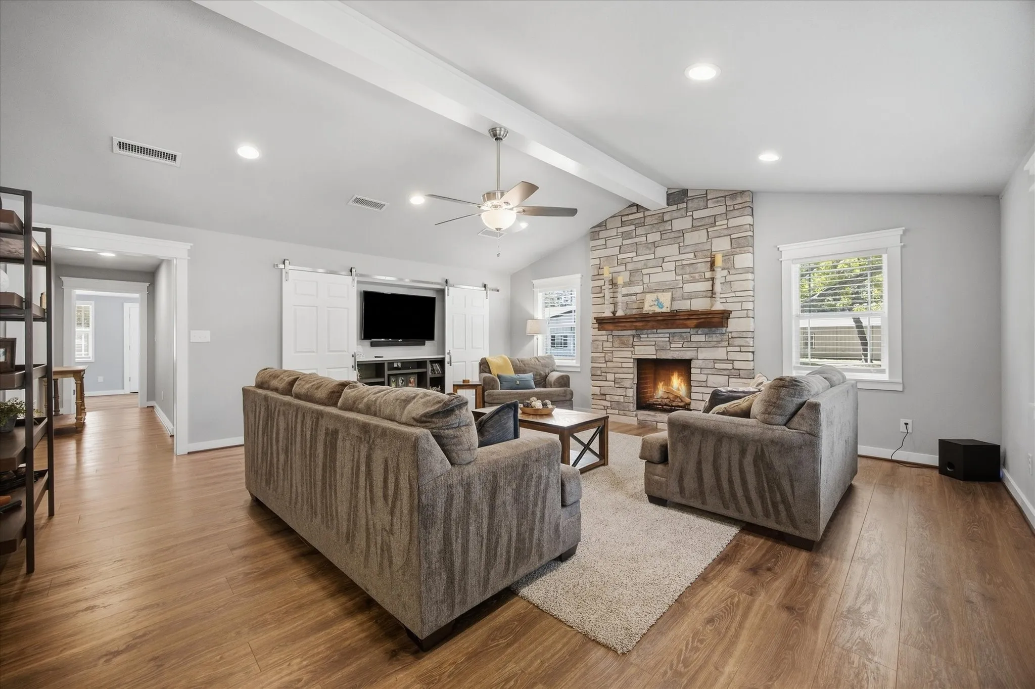 Vaulted ceiling with exposed beam, stone-clad fireplace with wood mantel, wood-finish flooring, recessed lighting, and barn door media cabinet