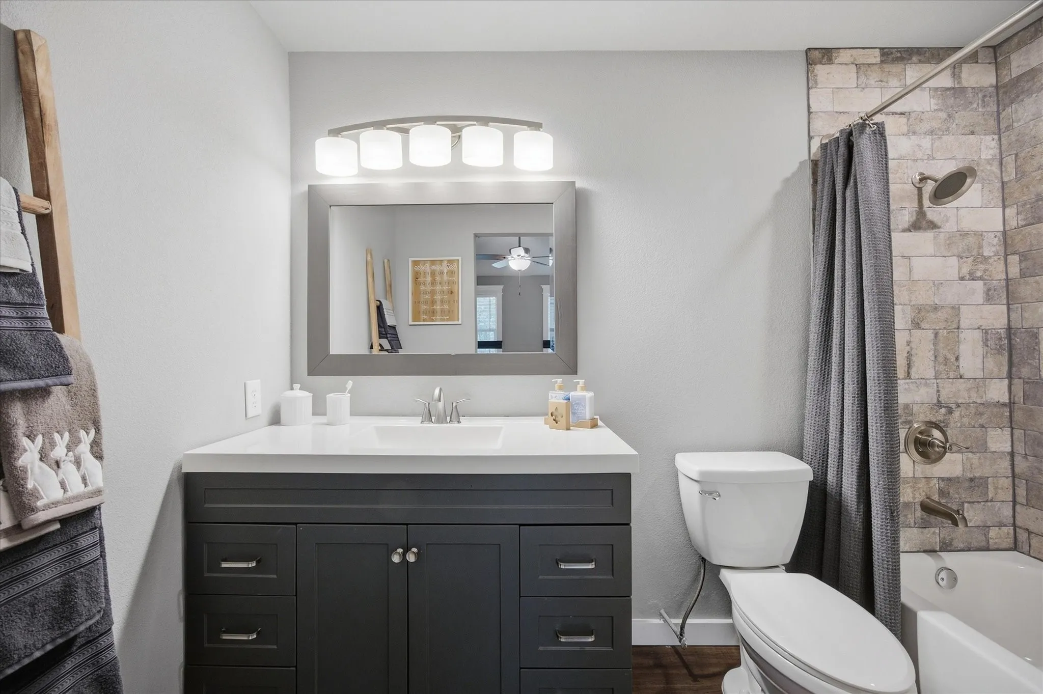 Bathroom featuring a dark gray vanity with a white countertop, a framed mirror, a four-light vanity fixture, and a tiled shower surround