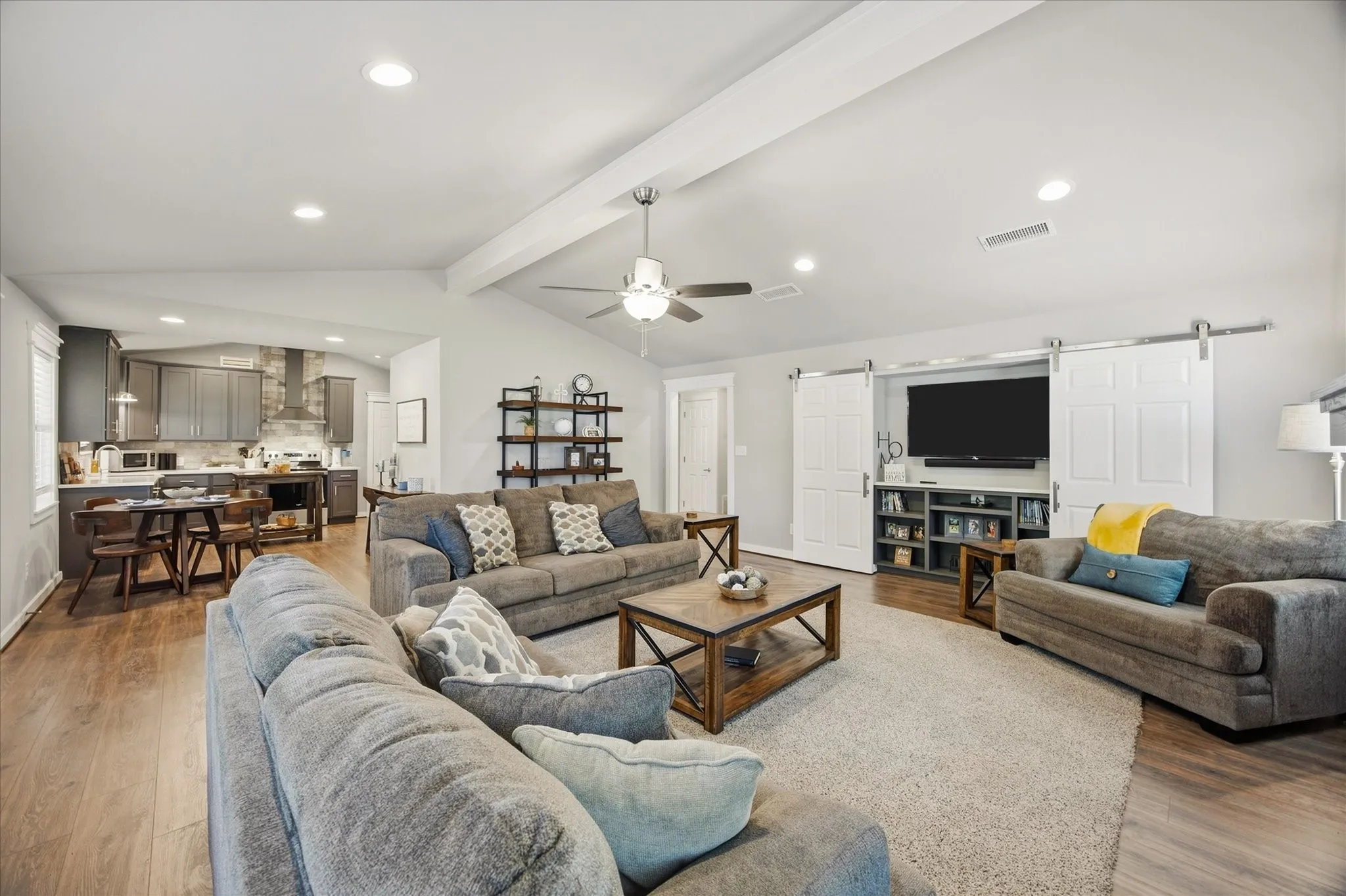 Vaulted ceiling with exposed beam, recessed lighting, wood-finish flooring, built-in entertainment center with barn door covers, and kitchen featuring dark cabinetry and tiled backsplash