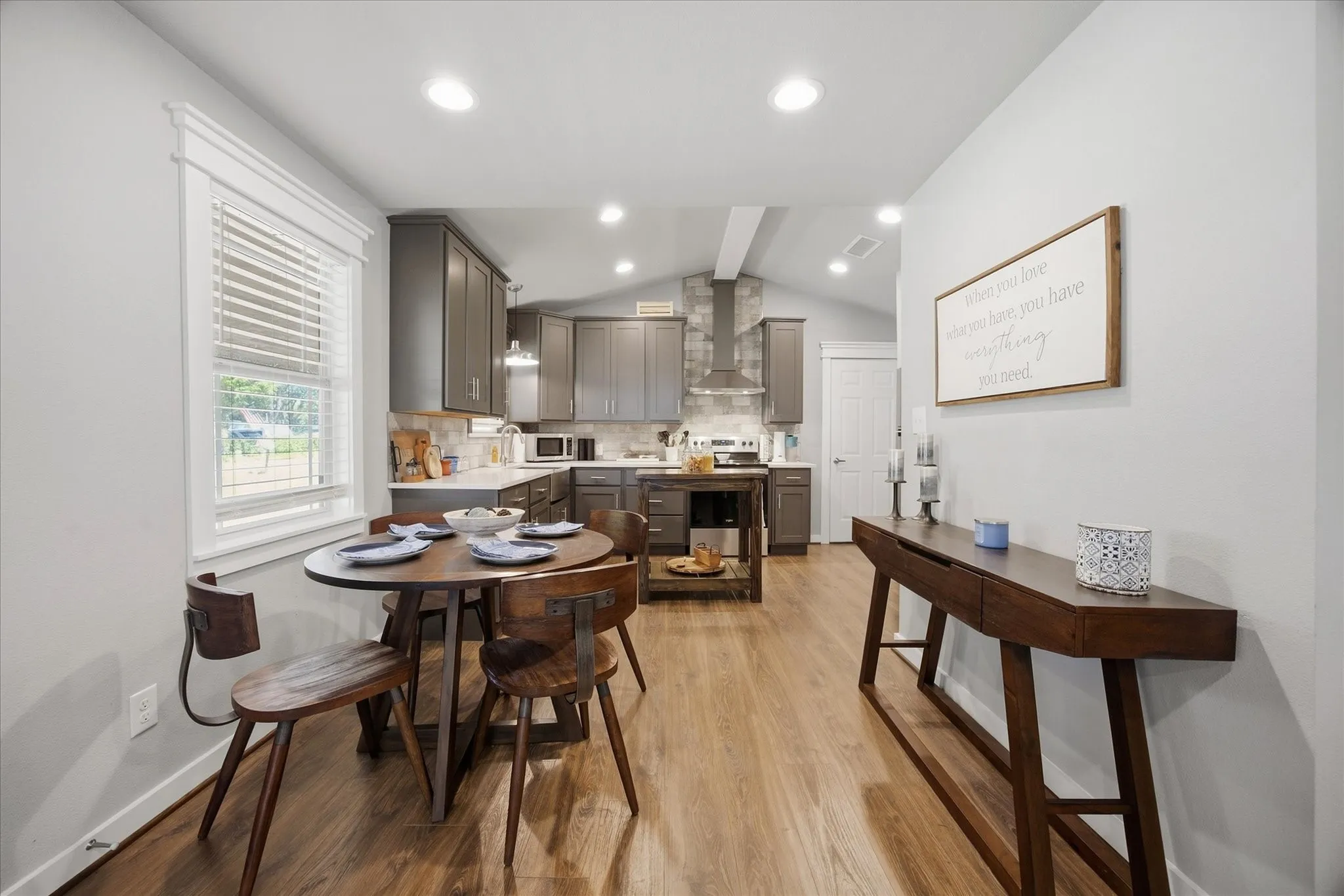 Open-concept living area featuring wood-finish flooring, recessed lighting, white window blinds, a kitchen with grey cabinetry, and a vaulted ceiling