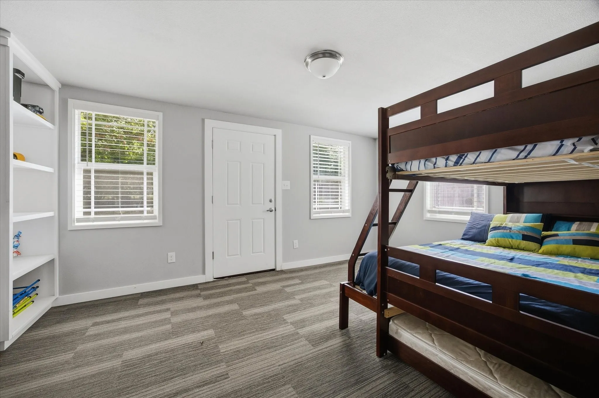 Light gray walls with integrated white shelving, two windows with white blinds, a white paneled door, and patterned carpet flooring
