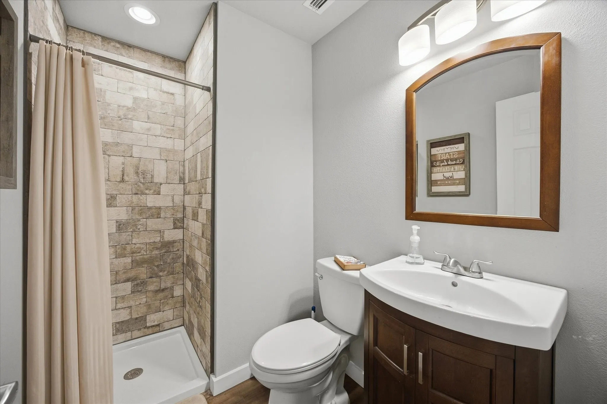 Shower with stone-look tile surround, wood-finish vanity with integrated sink, and wall-mounted light fixture