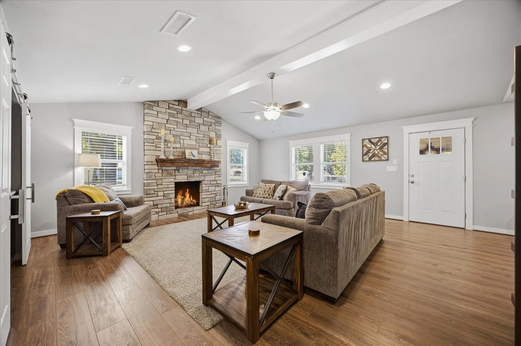 Living area featuring a stone-clad fireplace with a wood mantel, vaulted ceiling with exposed beam, wood-finish flooring, recessed lighting, and a white barn door
