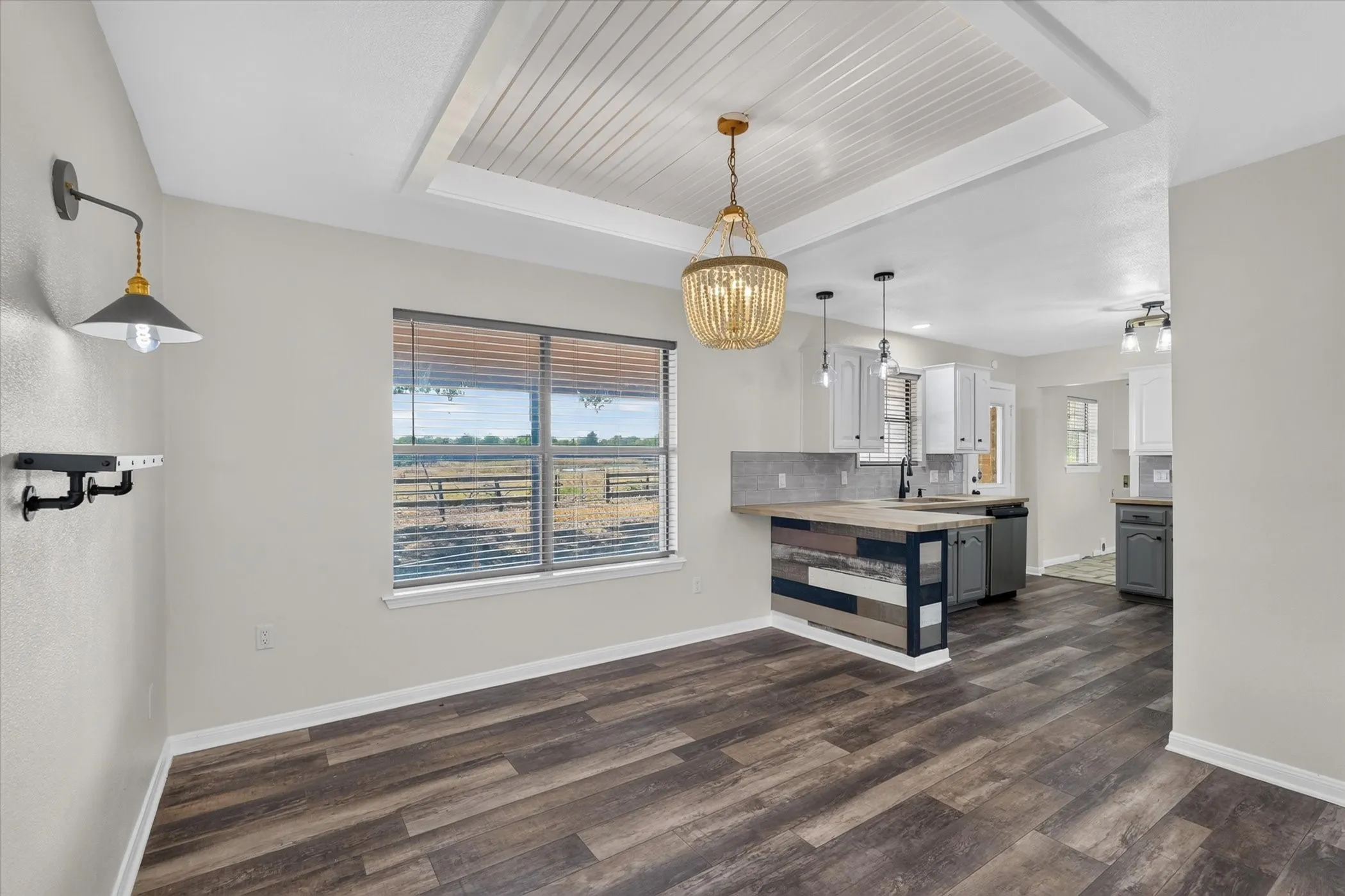 Wood-finish flooring, tray ceiling with beadboard detail, window with horizontal blinds, decorative wall sconce, and a kitchen island with a wood countertop