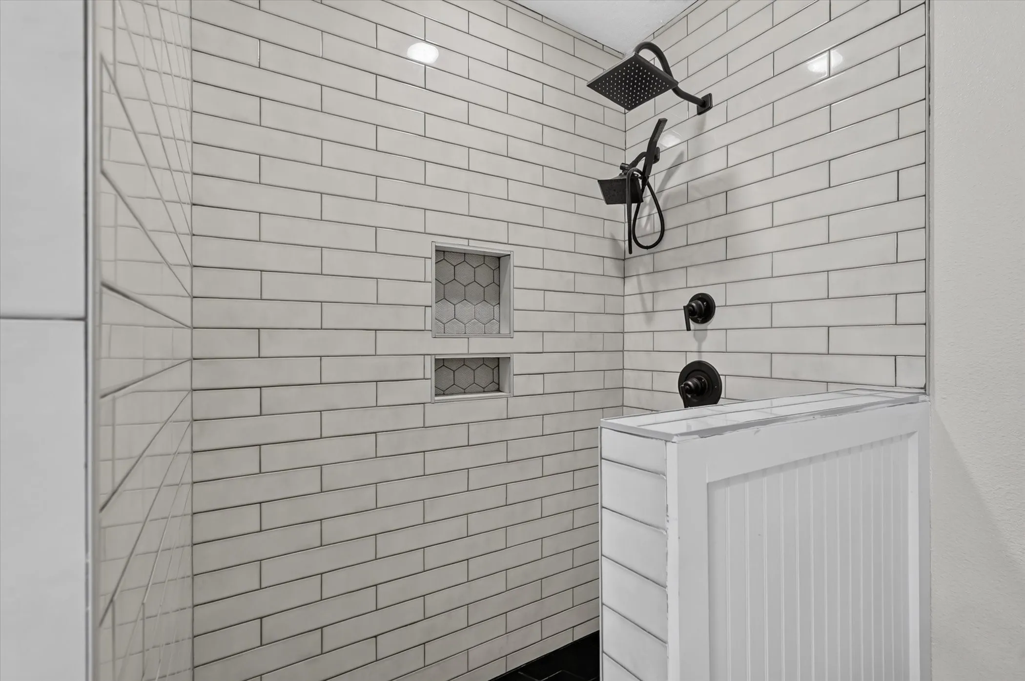 Shower enclosure with subway tile surround, two recessed niches with hexagonal tile, black rain shower head, and an additional handheld shower fixture