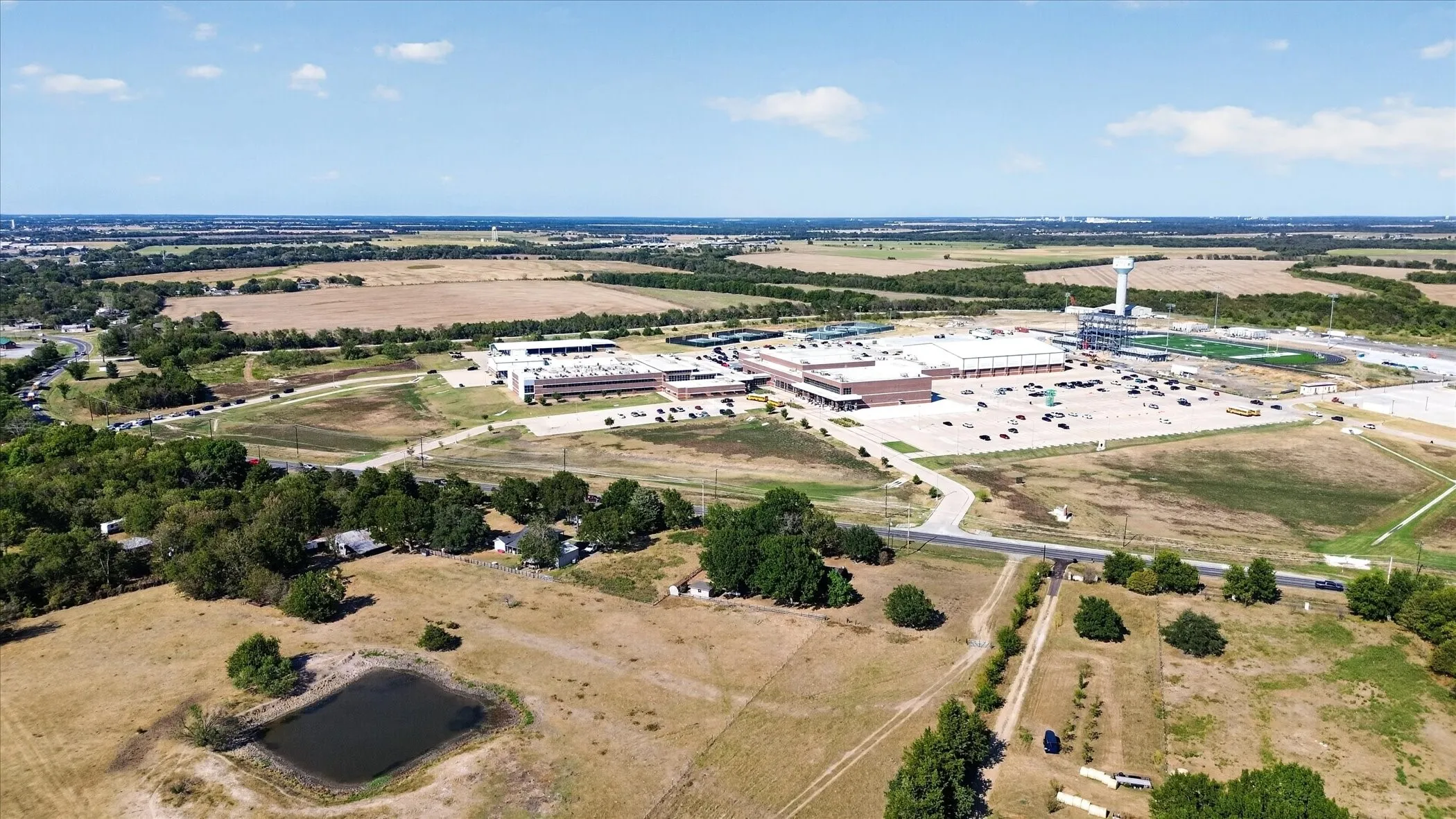 Expansive rural landscape featuring a pond, scattered trees, and distant commercial structures