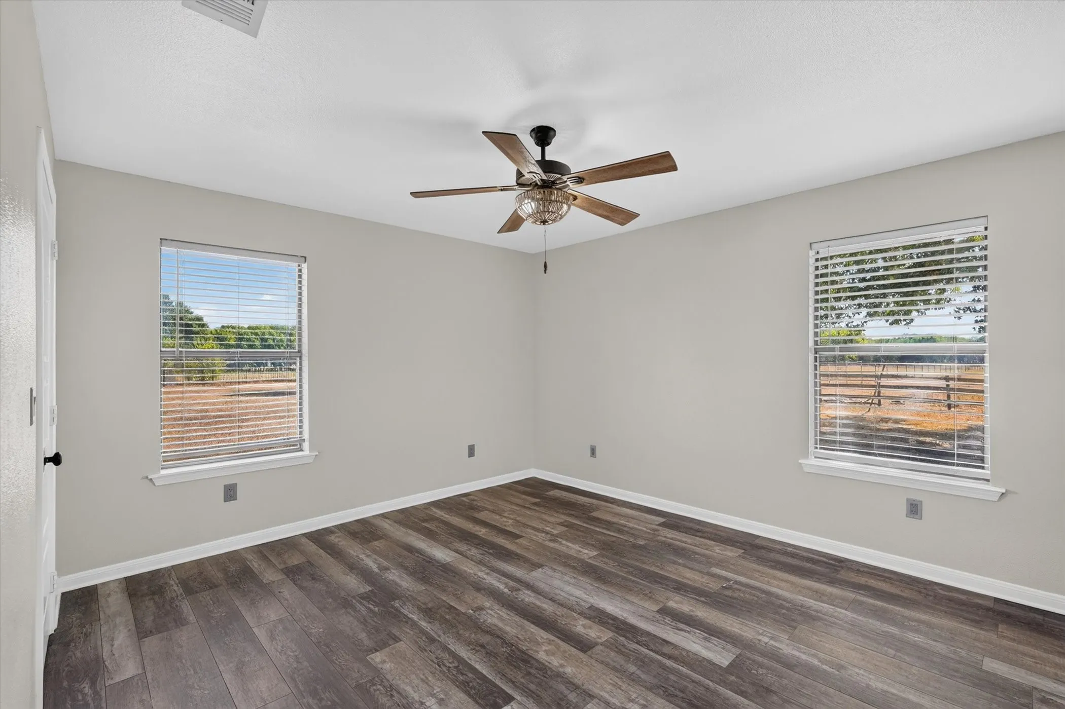 Warm wood-finish flooring, two windows with horizontal blinds, and a ceiling fan with light fixture
