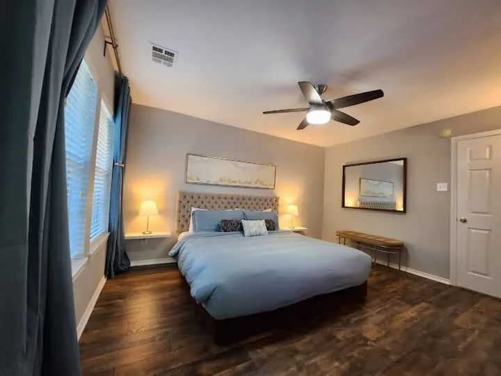 Wood-finish flooring, tufted headboard, ceiling fan with integrated lighting, wall-mounted floating shelves, and a paneled door
