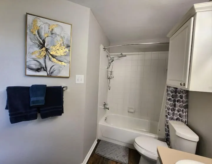Bathtub with tiled shower surround, commode with built-in overhead cabinetry, and wood-finish flooring