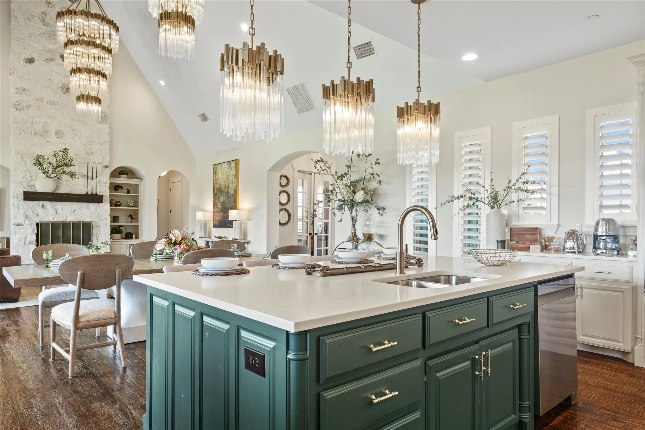 Vaulted ceiling, stone fireplace with wood mantel, large kitchen island with integrated sink, built-in shelving, and wood-finish flooring