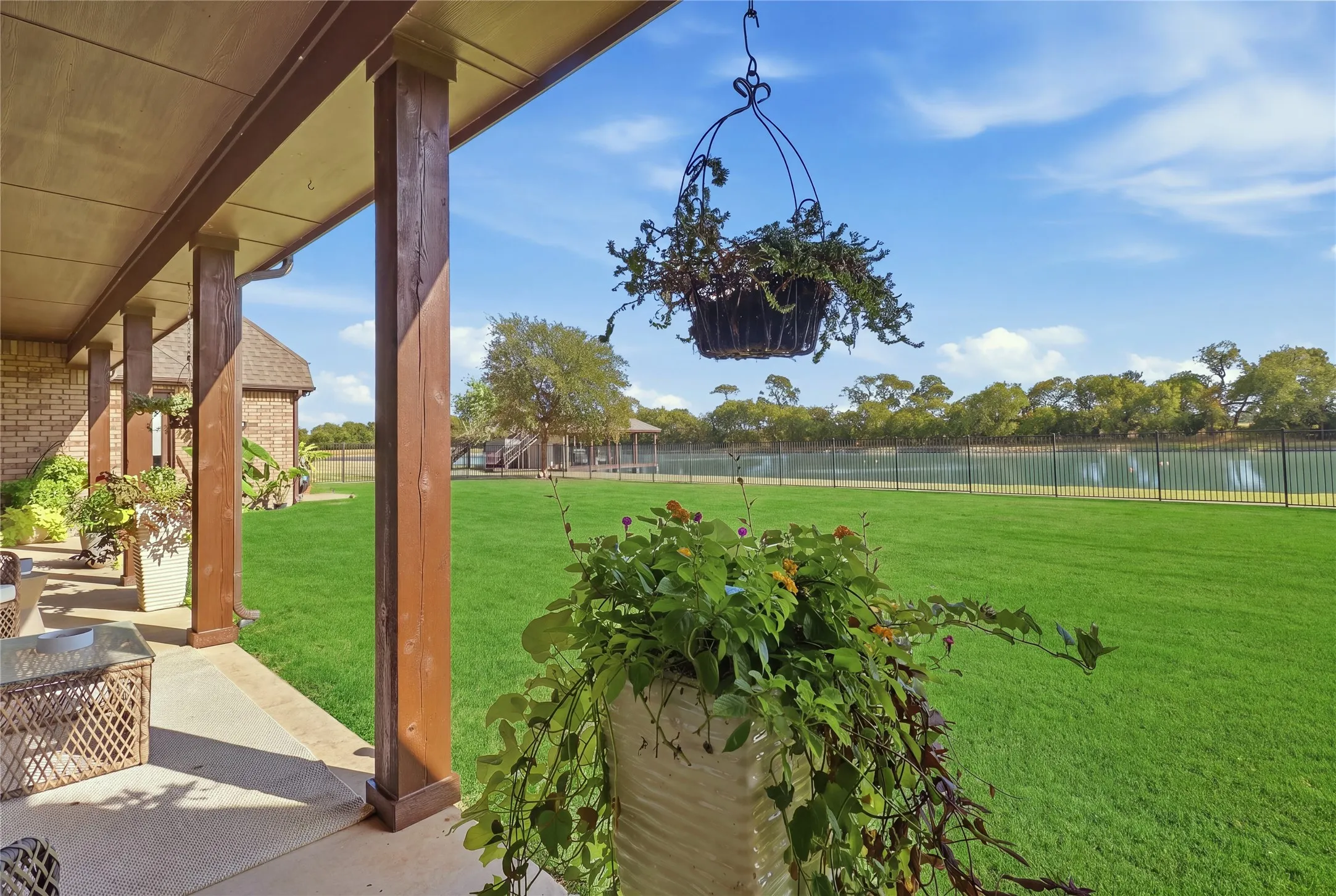 Covered patio with wood-finish support columns, brick exterior, expansive lawn, black metal fencing, and water feature beyond