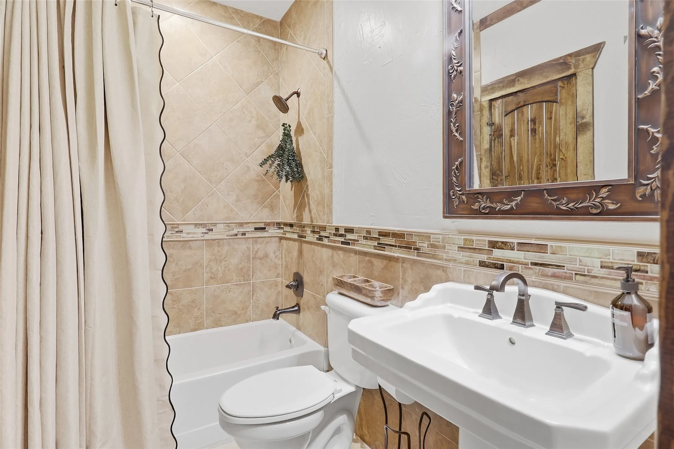 Pedestal sink with bronze-finish faucet, framed mirror, tiled wainscoting with mosaic accent, and a shower-over-tub with tiled surround