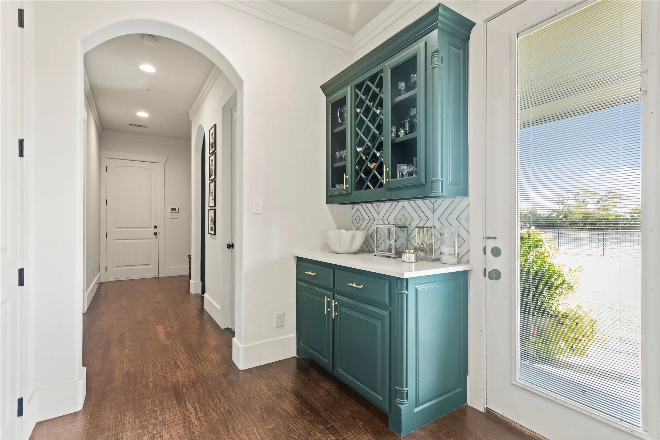 Built-in cabinetry with glass-front uppers and wine storage, white countertop, patterned tile backsplash, wood-finish flooring, and an arched hallway entry
