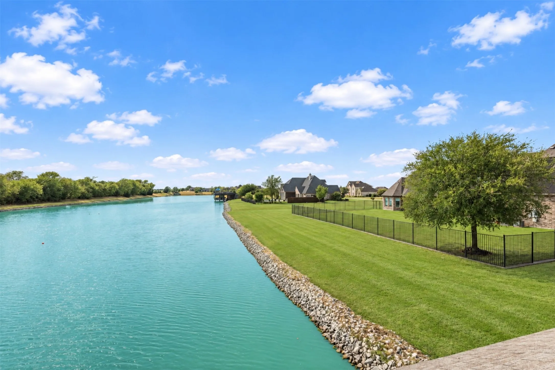 Waterfront property featuring a black metal fence, extensive green lawn, and mature trees