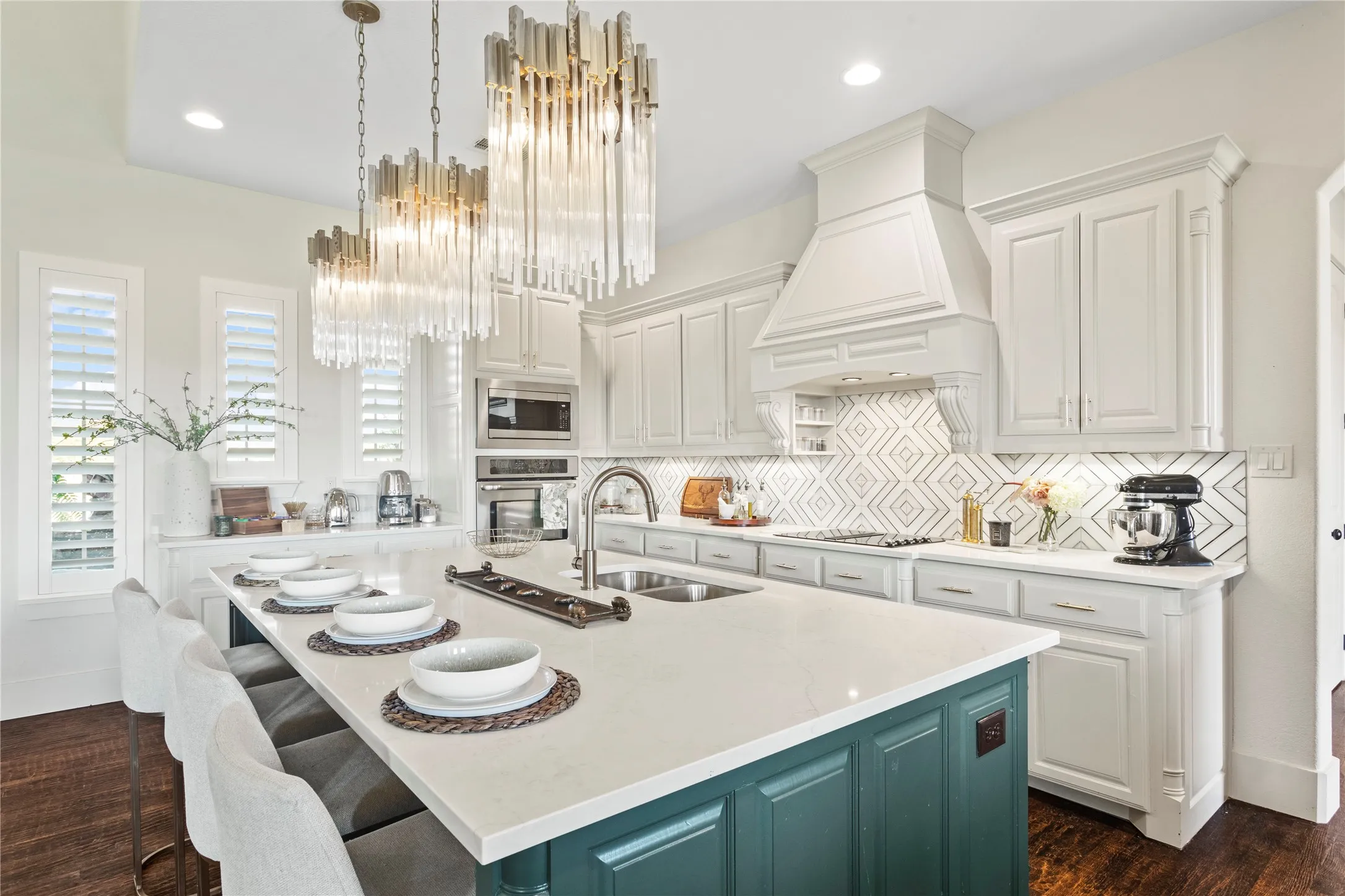 Kitchen with a teal island, white perimeter cabinetry, a white range hood, a patterned tile backsplash, and three tiered glass and brass chandeliers