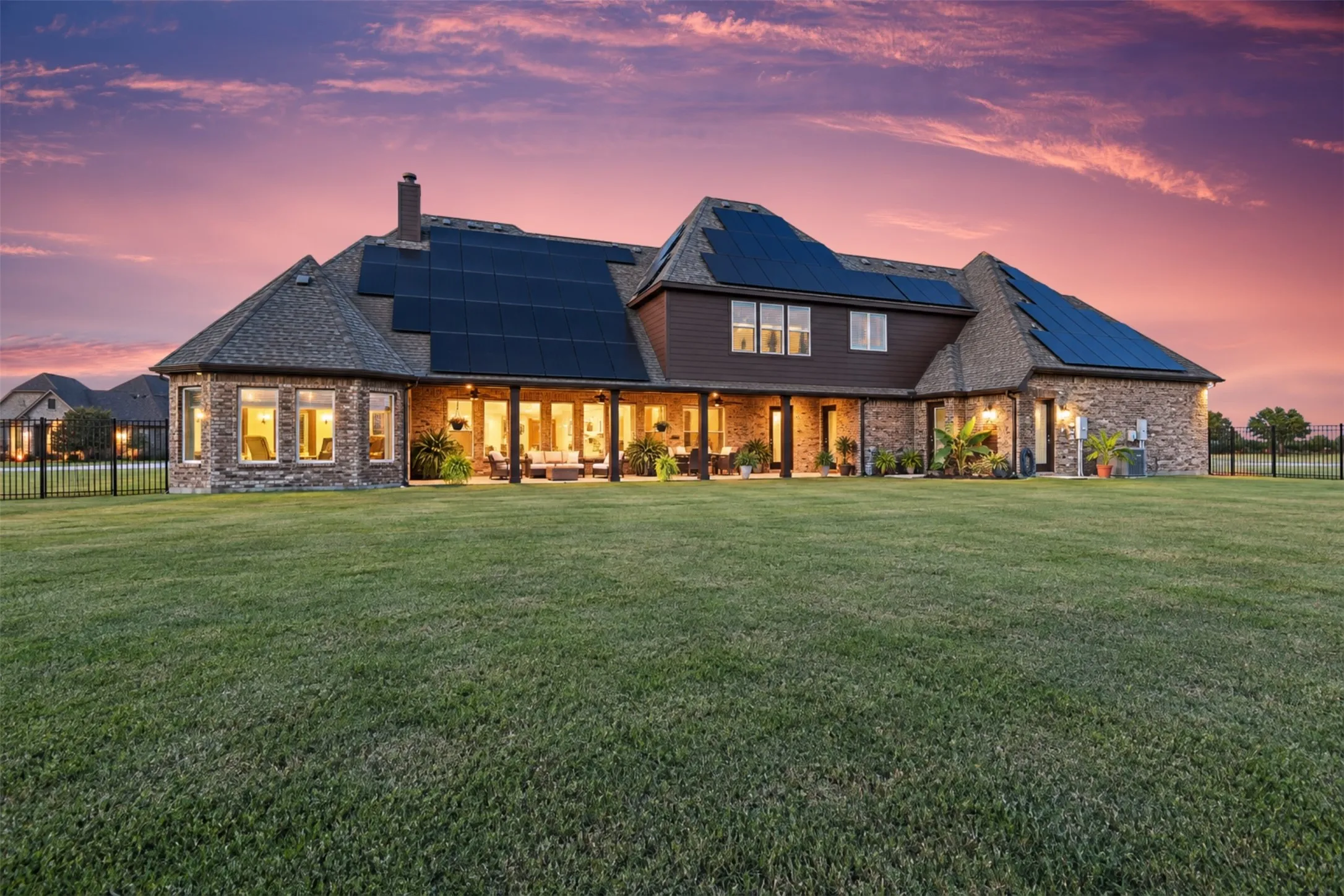 Expansive rear facade featuring brick and siding, multiple solar panel arrays, a covered patio, and a large grassy yard enclosed by black fencing