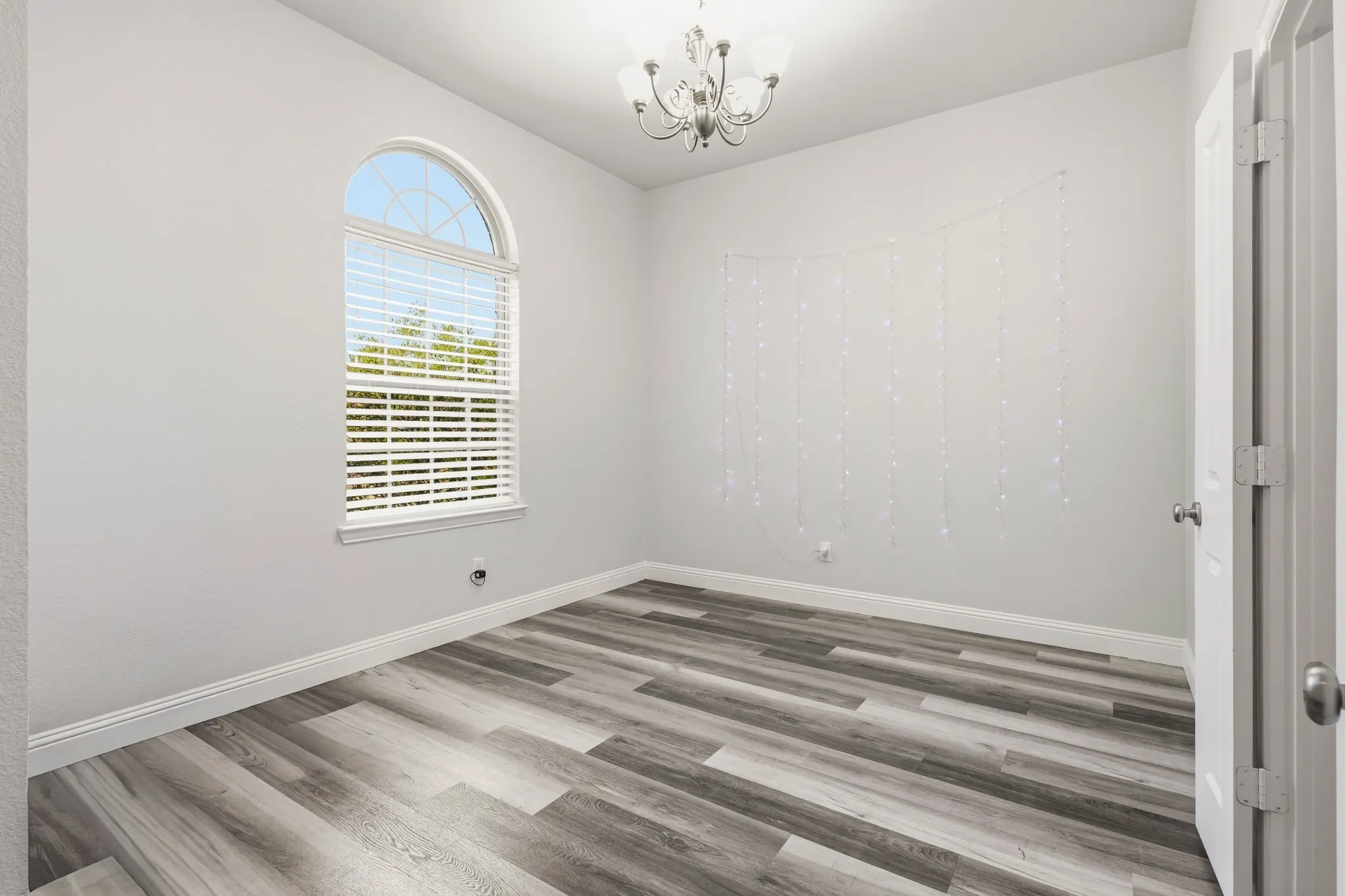 Arched window with blinds, wood-finish flooring, white walls, and a ceiling-mounted chandelier