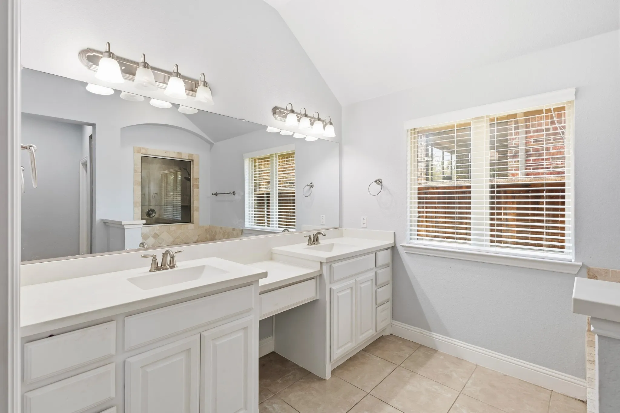 Dual vanity with white cabinetry and integrated sinks, vaulted ceiling, large wall-mounted mirror, and tiled flooring