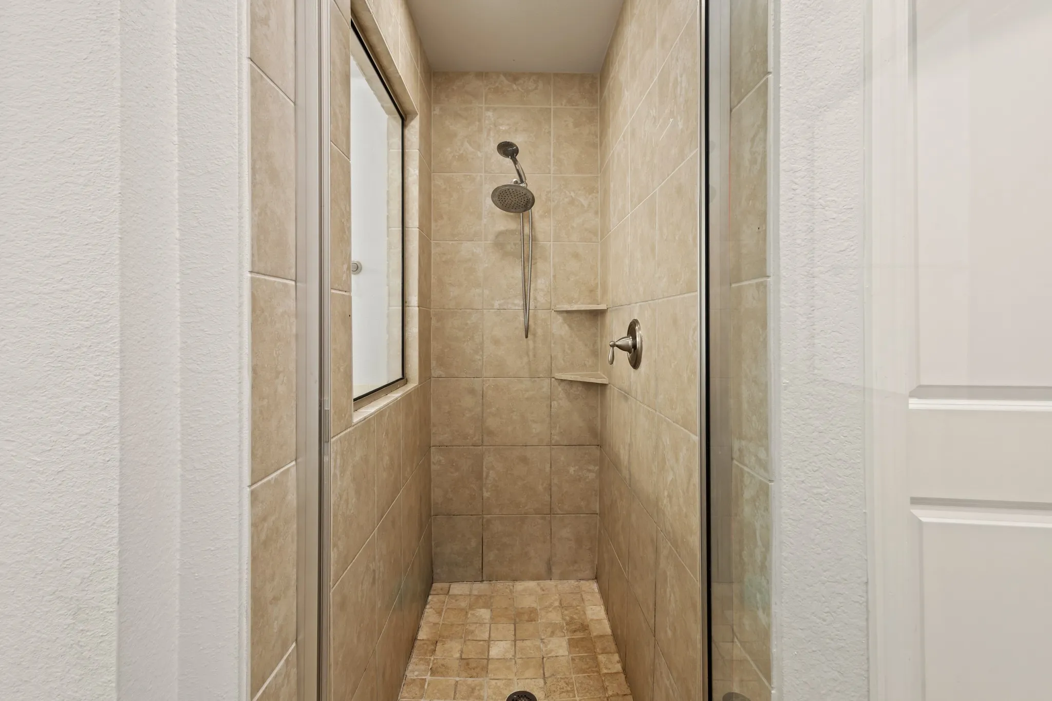 Tiled shower enclosure featuring a fixed showerhead, built-in shelving, and a glass door