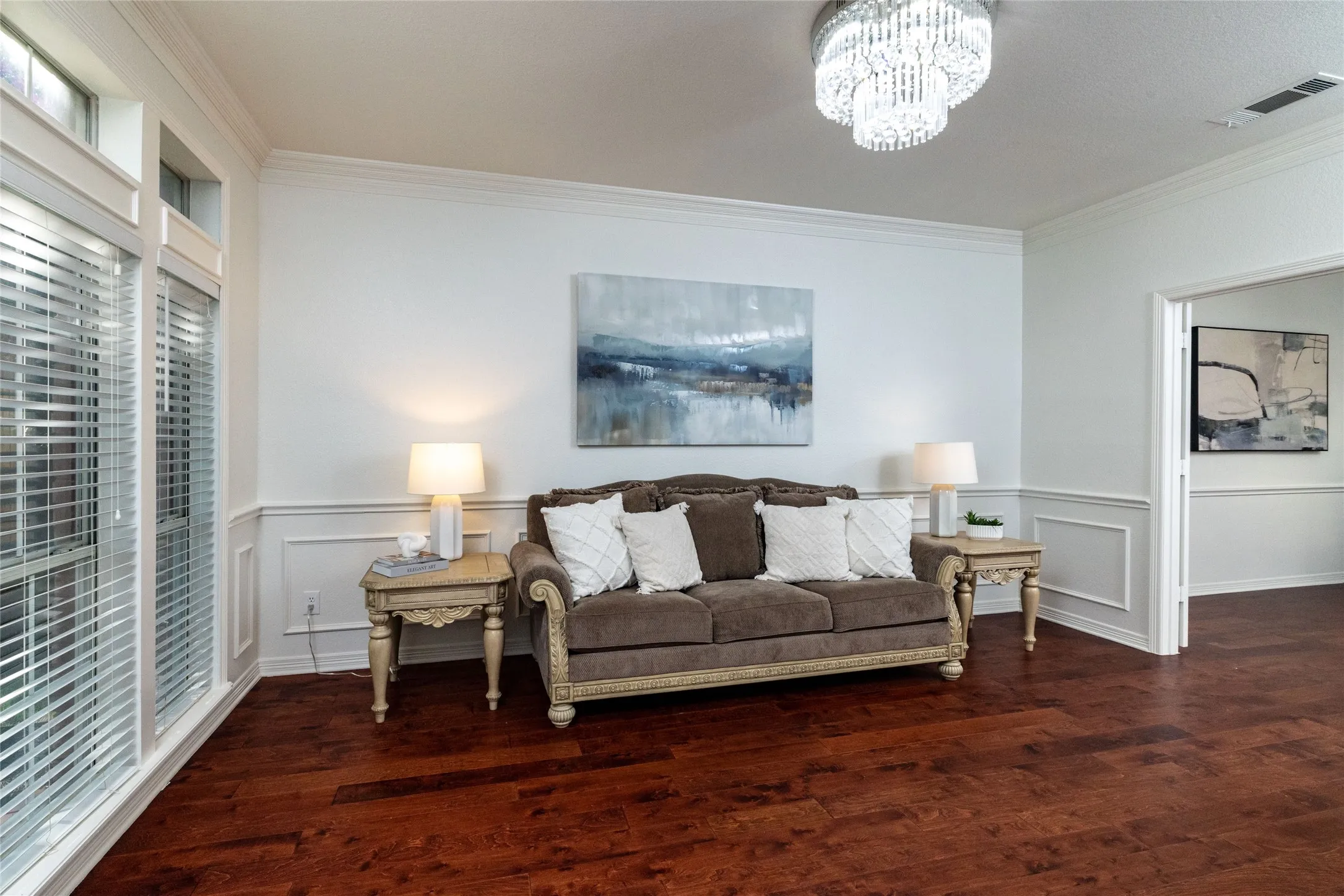 Rich wood-finish flooring, elegant crown molding, decorative wainscoting, large windows with blinds, and a tiered crystal chandelier
