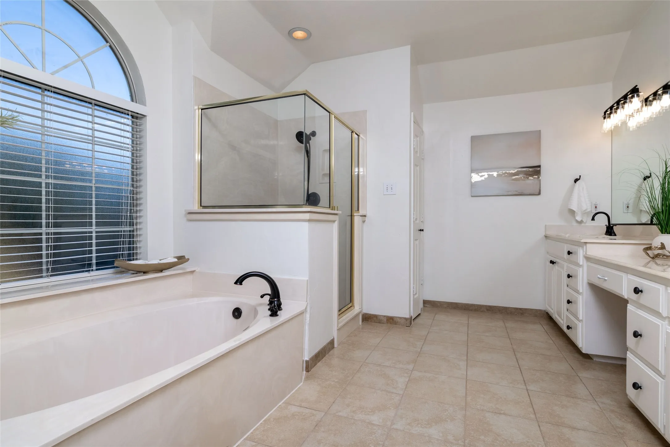 Arched window above a garden tub with matte black fixtures, an enclosed shower, dual vanity with white cabinetry, and tile flooring