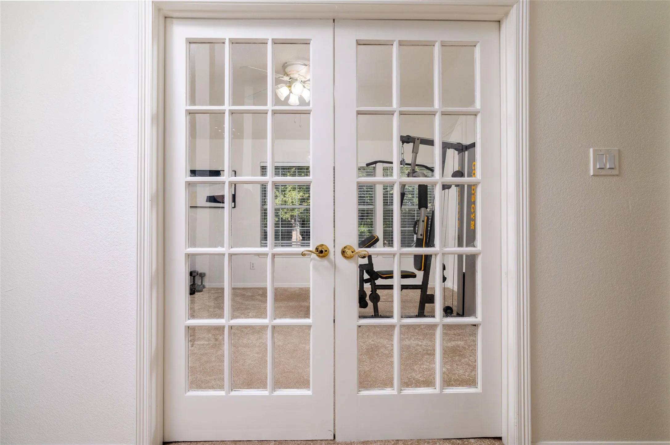 Double French doors with brass hardware opening into a carpeted room featuring a ceiling fan and natural light