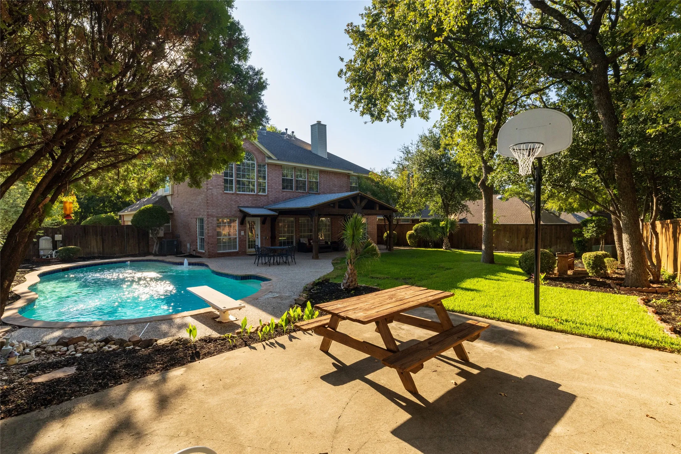Expansive backyard featuring a swimming pool with diving board, covered patio, mature trees, and a basketball hoop