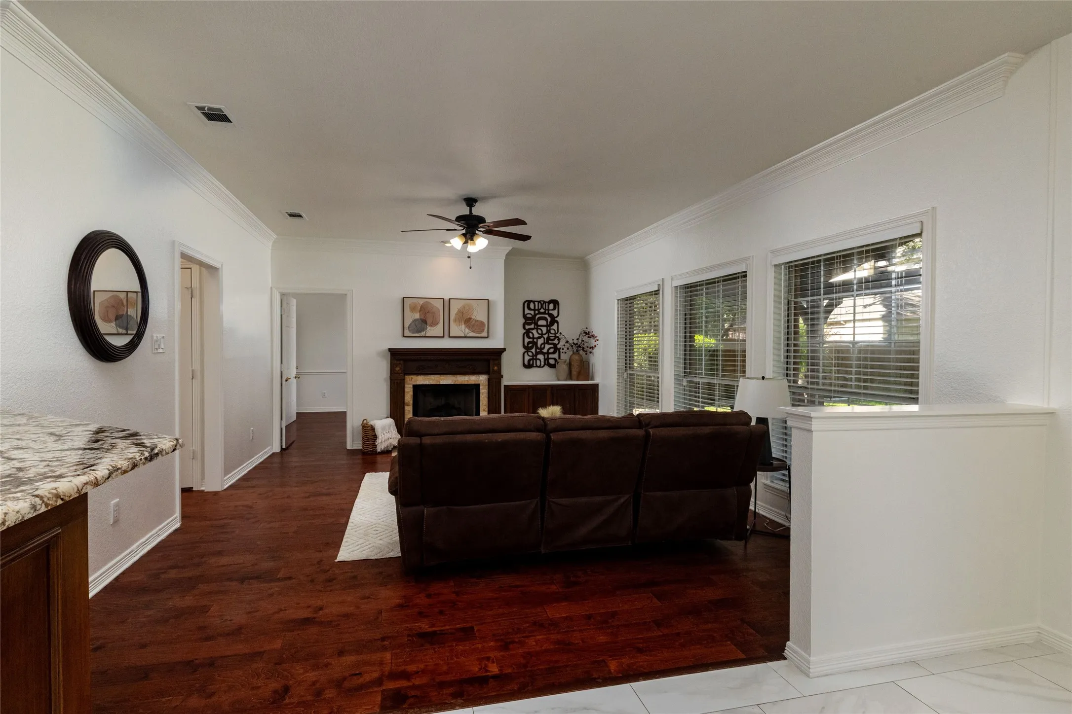 Hardwood-finish flooring, crown molding, a fireplace with a wood mantel, a ceiling fan, and multiple windows with blinds
