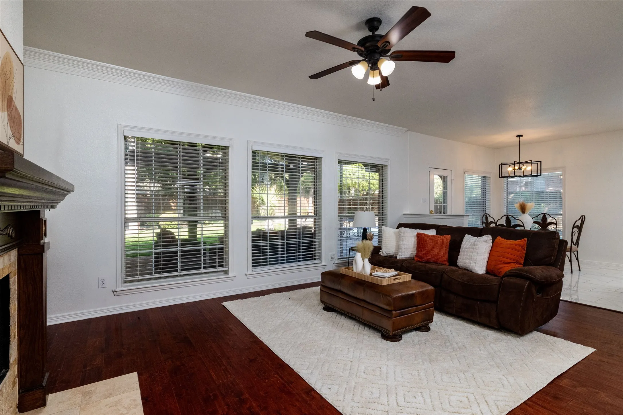 Wood-finish flooring, crown molding, a built-in fireplace with a stone surround, multiple windows with blinds, and a ceiling fan with integrated lighting