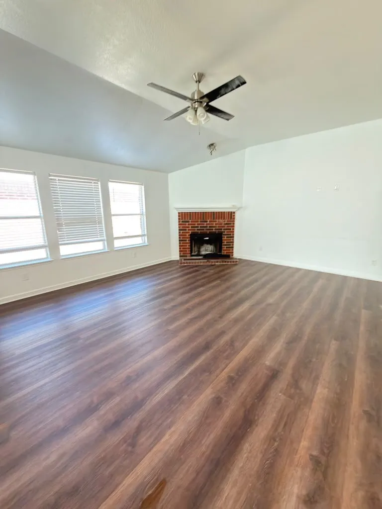 Wood-finish flooring, brick-surround fireplace, vaulted ceiling, multiple windows with blinds, and ceiling fan with integrated lighting