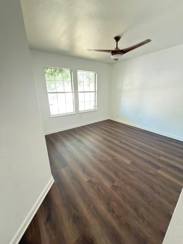 Wood-finish flooring, two large windows, white walls, and a ceiling fan with integrated lighting