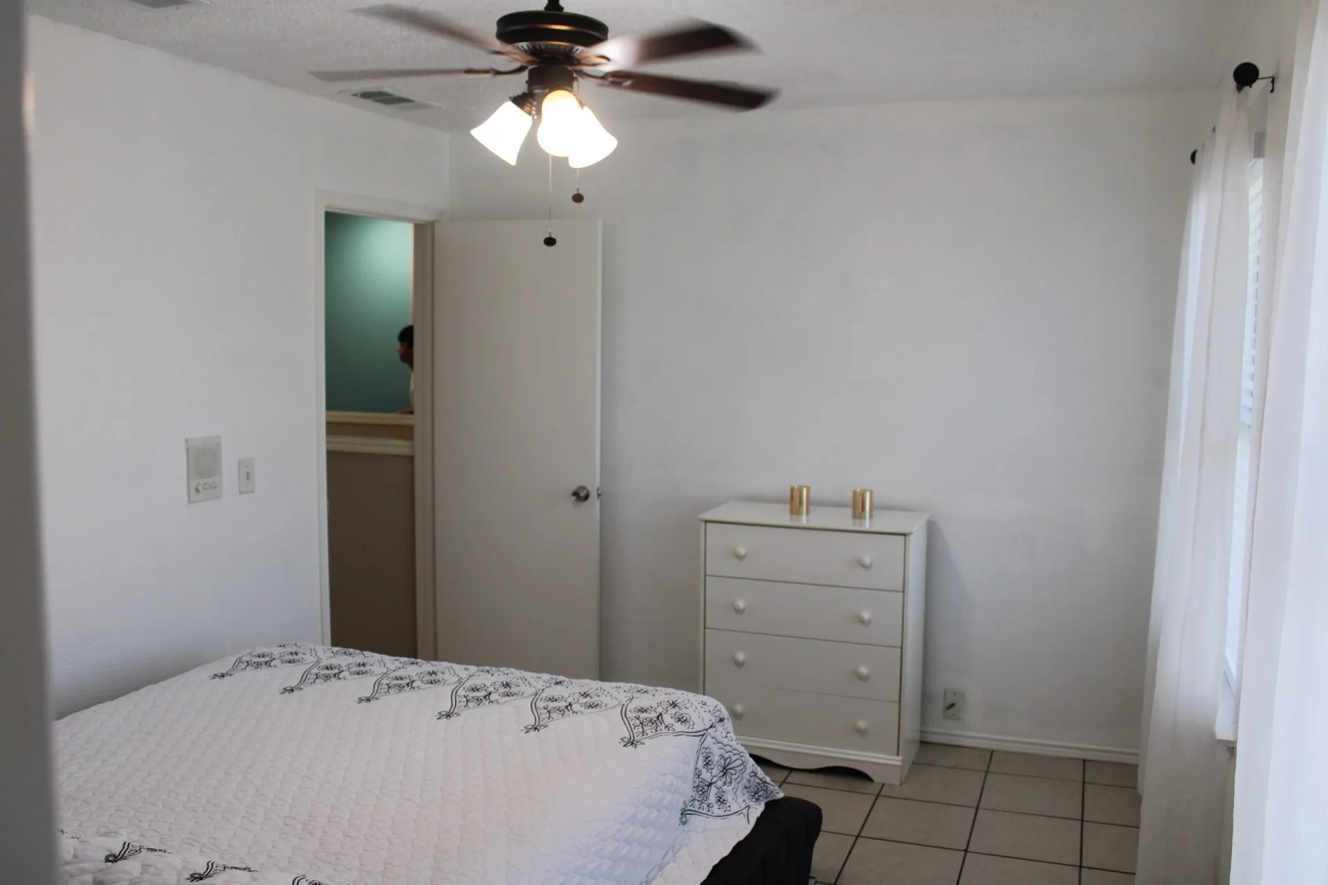Ceiling fan with integrated lighting, tiled flooring, white walls, and a window with curtain rod