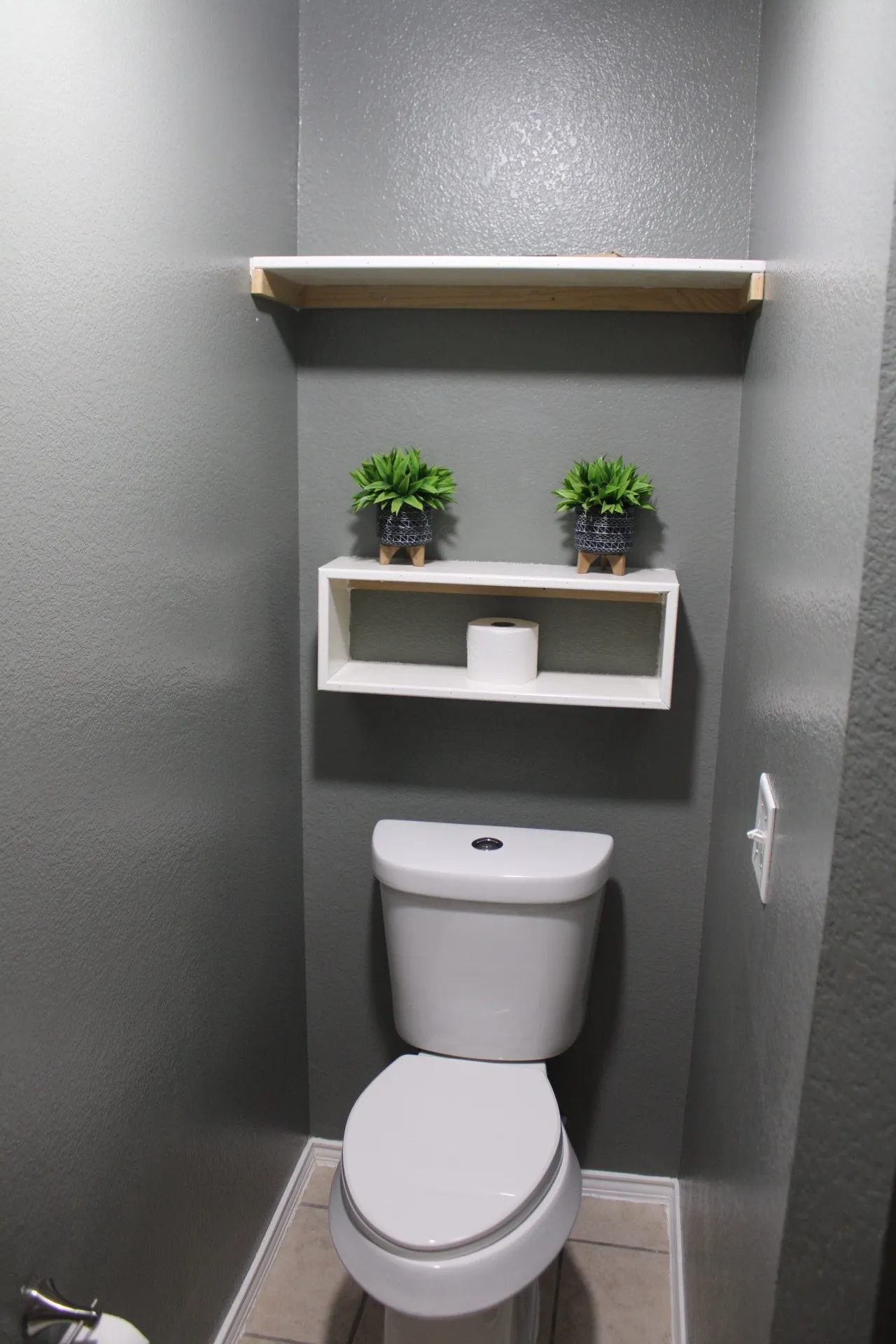 Recessed toilet alcove featuring a white commode, two wall-mounted shelves, and neutral-toned floor tile