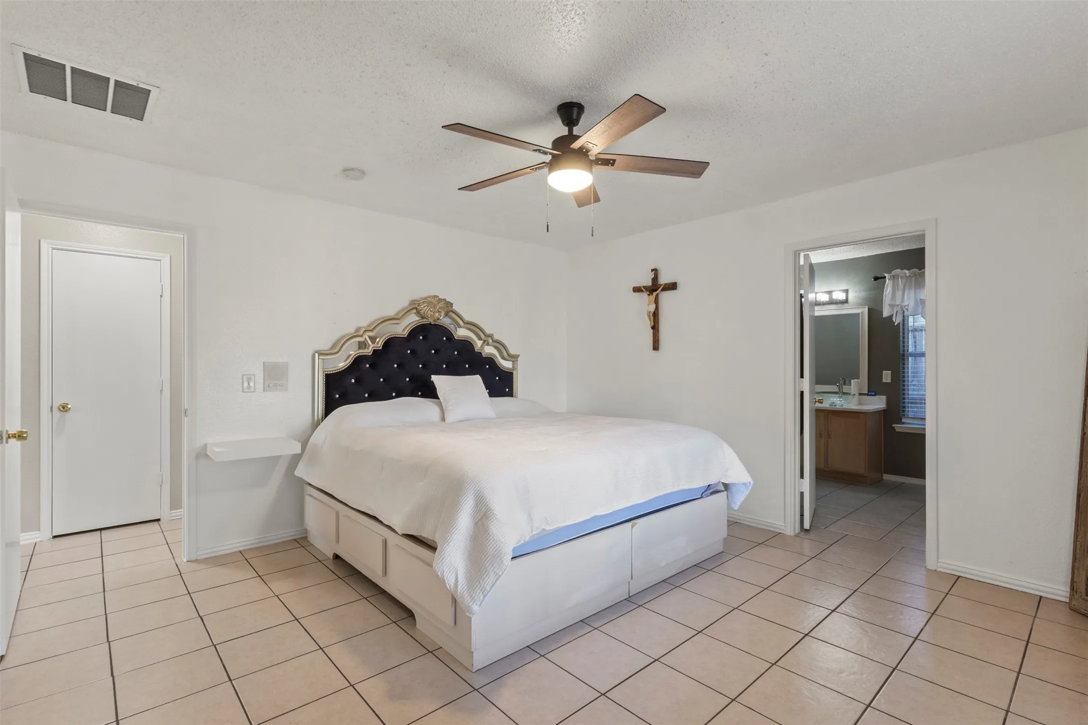 Tile flooring, ceiling fan with light fixture, white walls, built-in shelving, and a separate en-suite bathroom featuring a vanity with a wood-finish cabinet and dual sinks