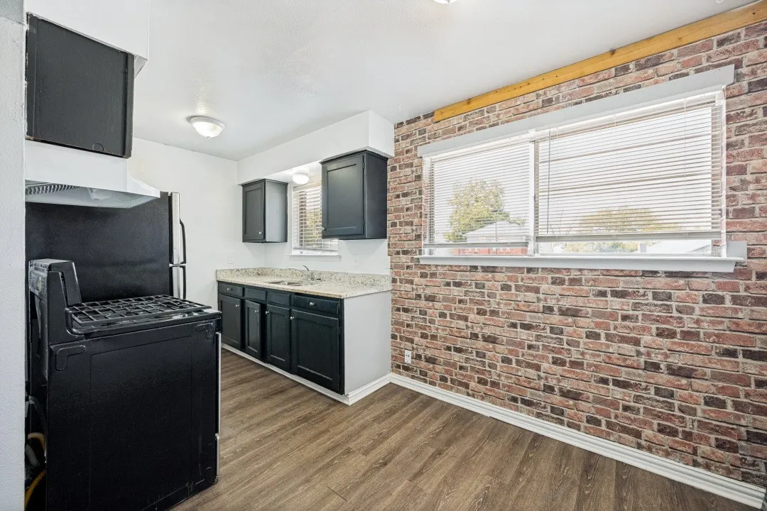 Kitchen featuring wood-finish flooring, dark cabinetry, light countertops, and a brick-finish accent wall