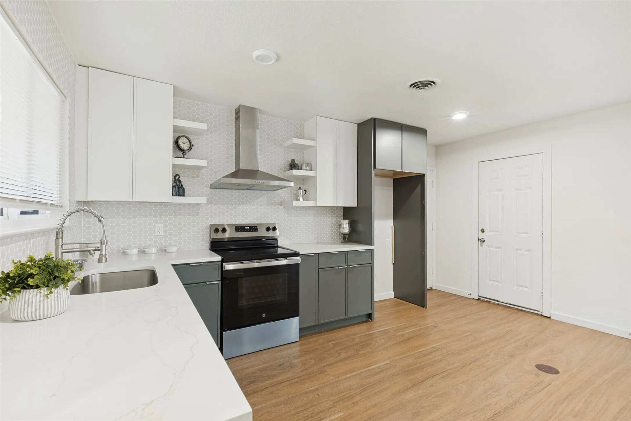 Modern kitchen featuring light wood-finish flooring, white countertops, two-tone cabinetry, a stainless steel range hood, and a geometric tile backsplash