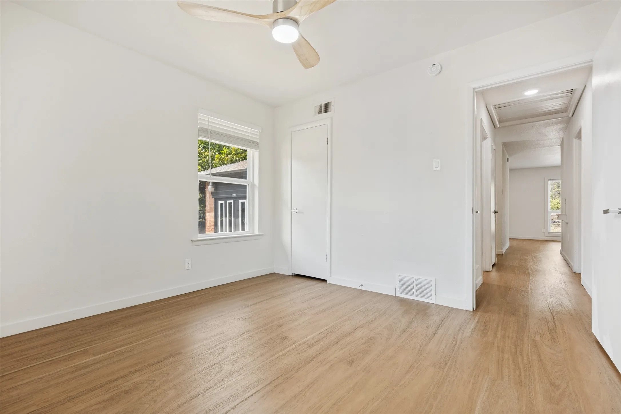 Wood-finish flooring throughout, bright white walls, window with blinds, modern ceiling fan with integrated lighting, and a solid core door