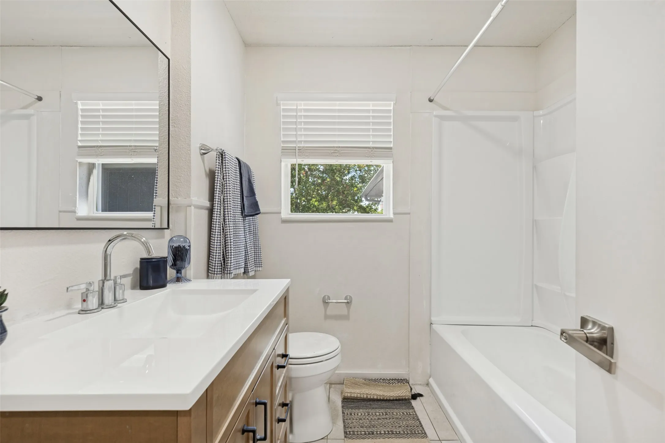 White bathroom featuring a single vanity with a white countertop and wood-finish cabinetry, a built-in tub with a white surround, and dual windows with blinds