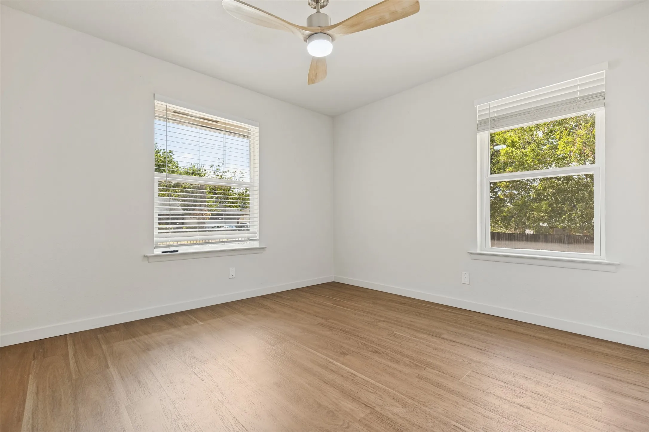 Neutral wall paint, wood-finish flooring, two windows with blinds, and a ceiling fan with integrated lighting