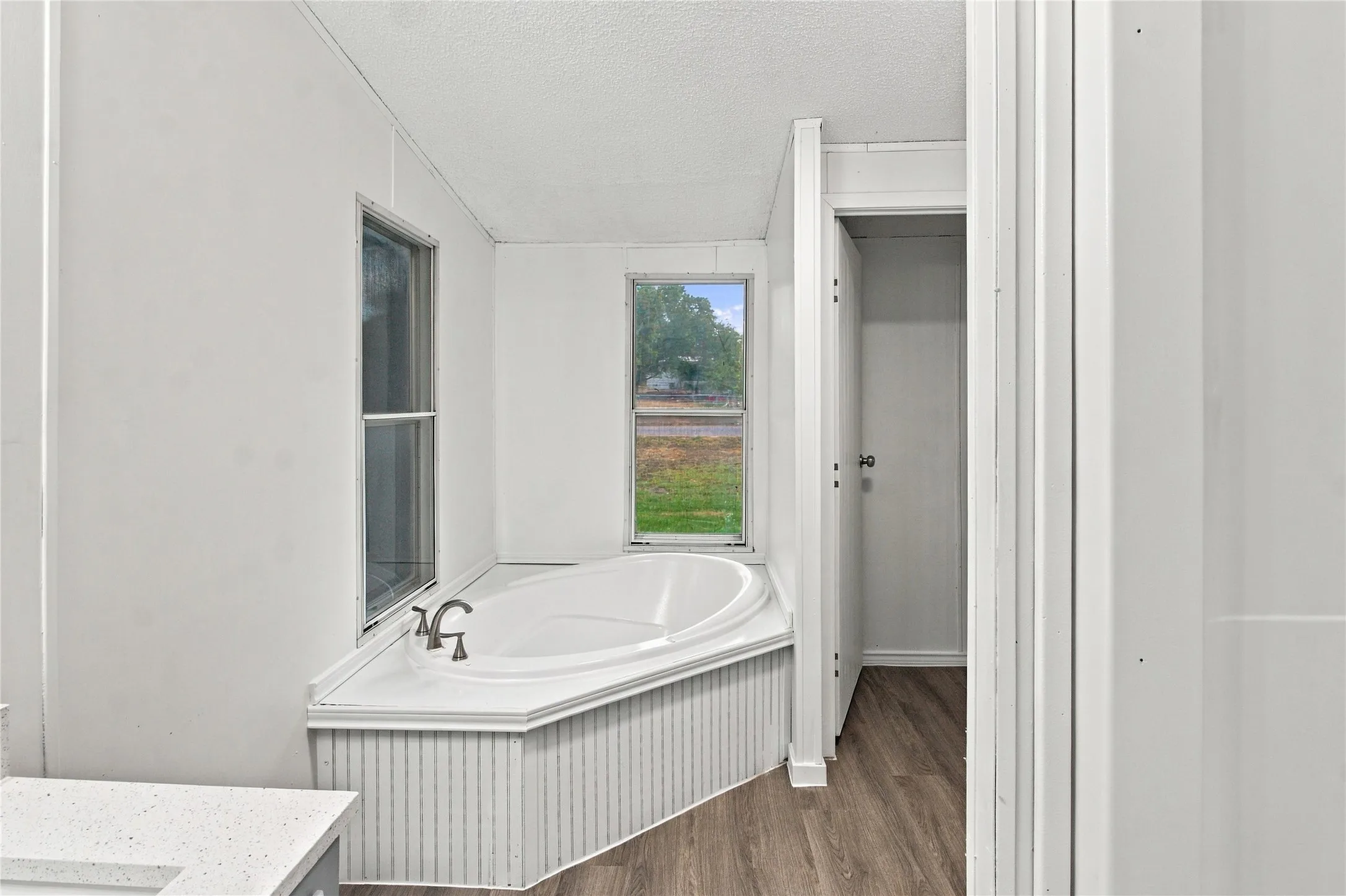 Corner soaking tub with integrated white paneled surround, set against two windows, featuring wood-finish flooring, and a white countertop