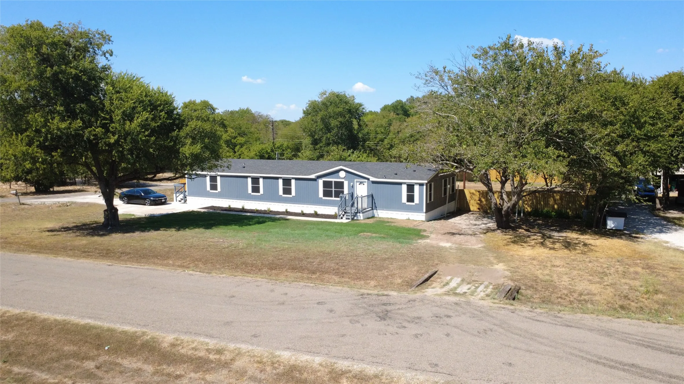 Blue exterior siding with white trim, dark shingle roof, front porch with railings, and mature trees on the property
