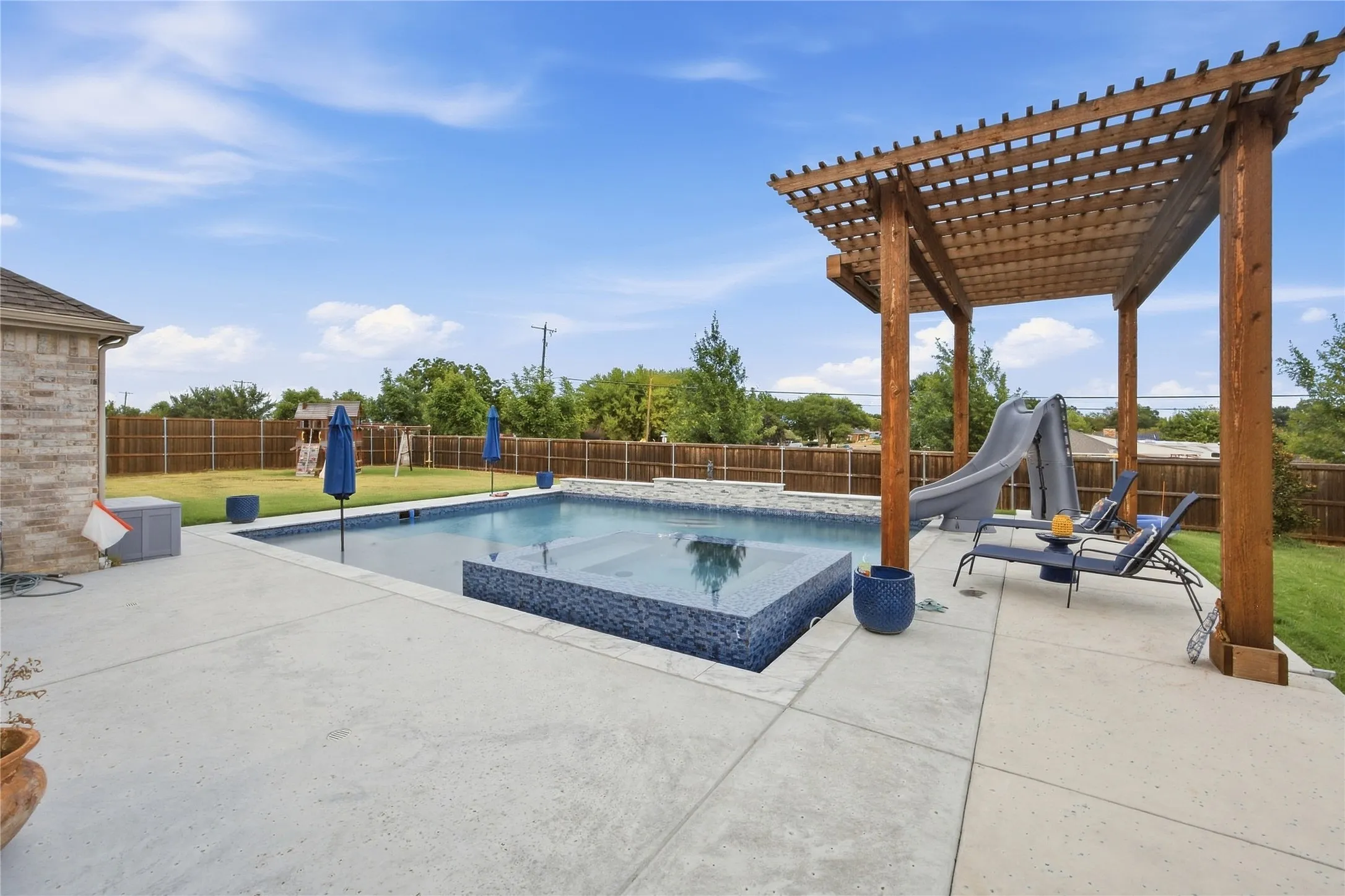 Rectangular pool with integrated spa and blue mosaic tile, wood pergola, and expansive concrete patio
