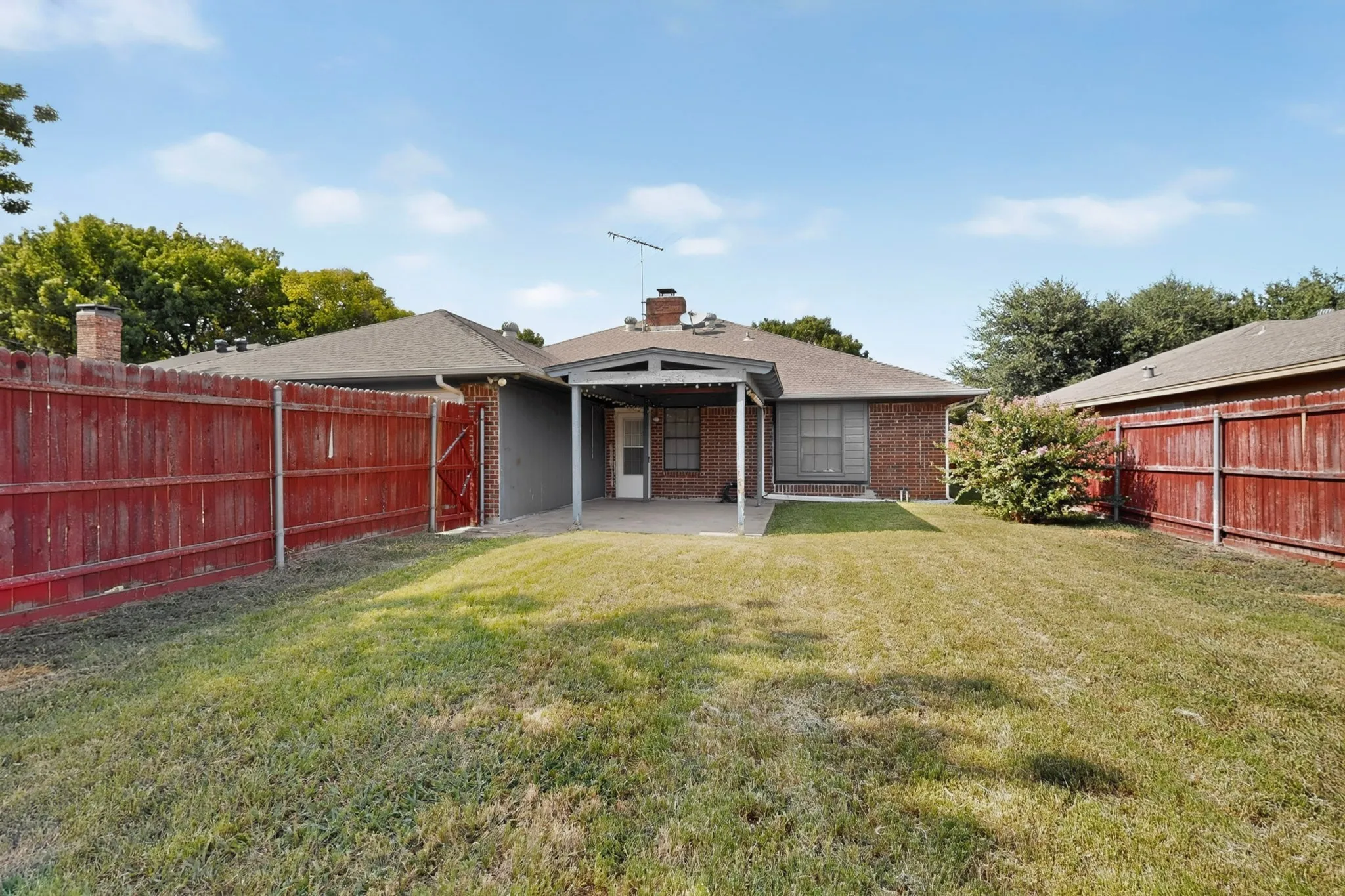 Expansive backyard with a covered patio, red-stained wood privacy fence, and brick exterior