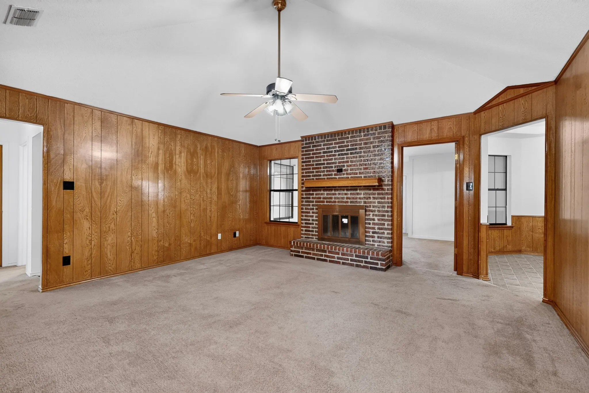 Vaulted ceiling with a ceiling fan, brick fireplace with a wood mantle, wood paneling, and carpet flooring