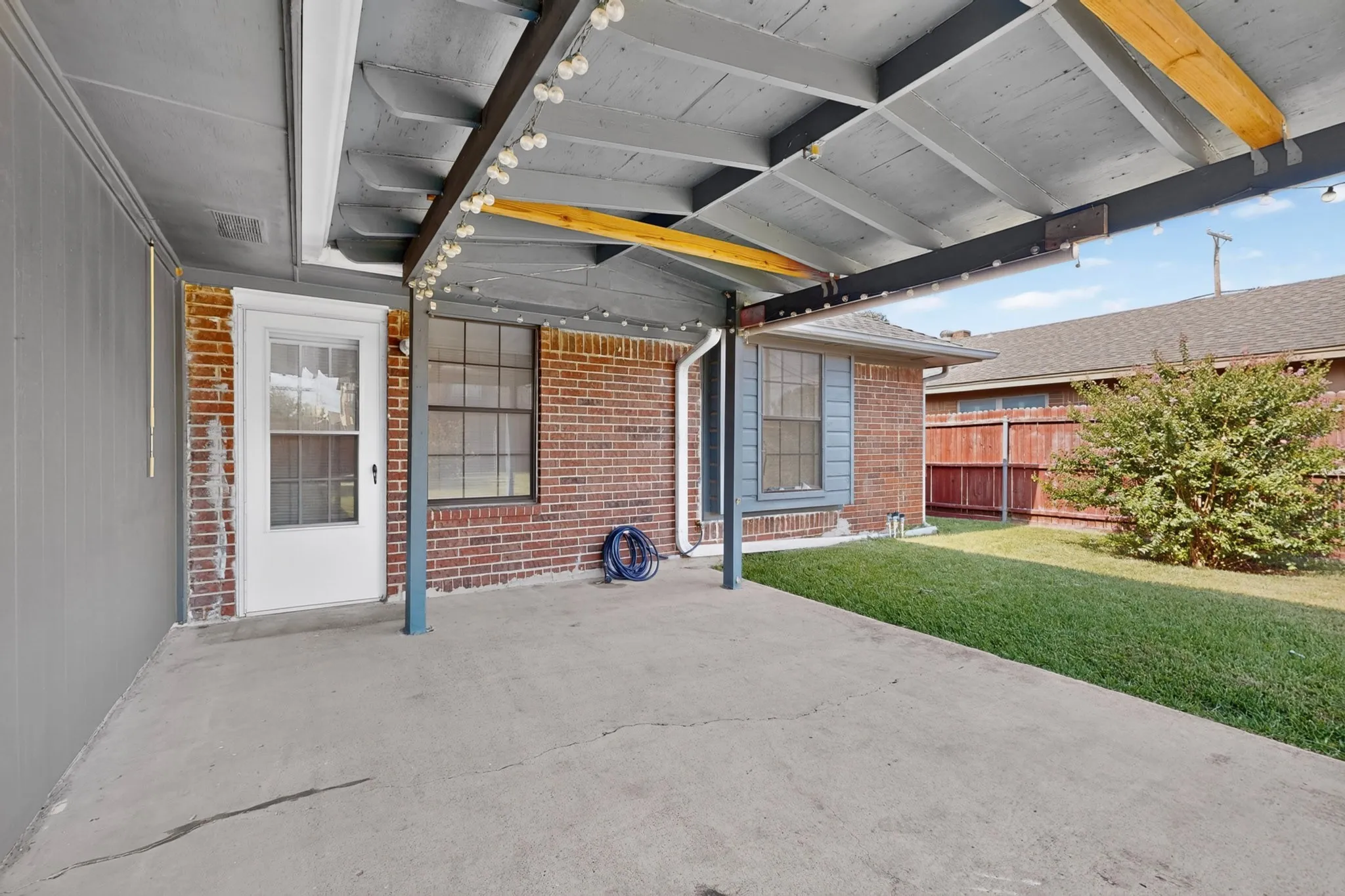 Covered patio with a concrete surface, supported by exposed wood beams, brick exterior, and a manicured lawn
