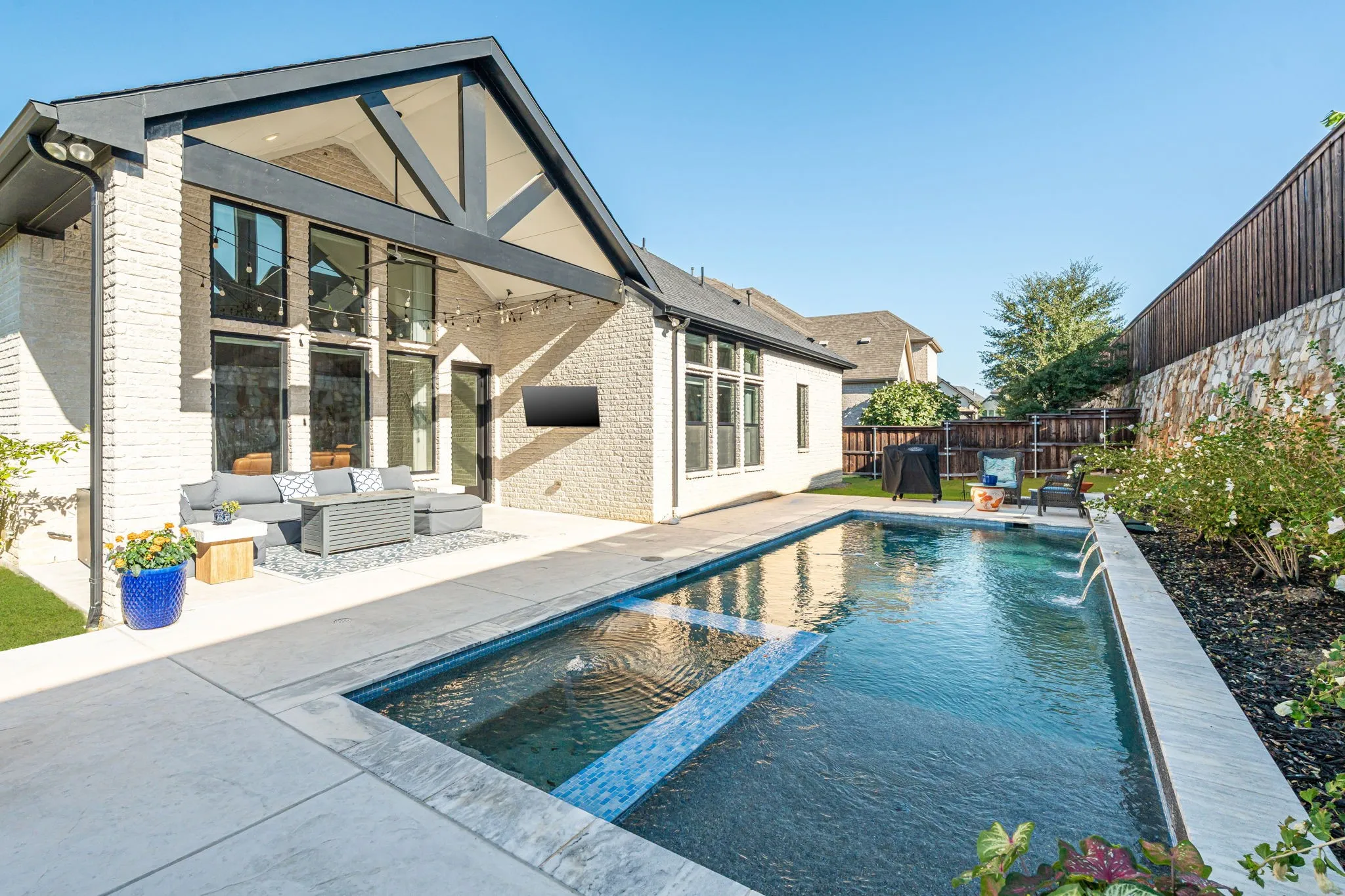 White brick exterior with black trim, a large outdoor patio, a rectangular swimming pool with water features, and a built-in outdoor television