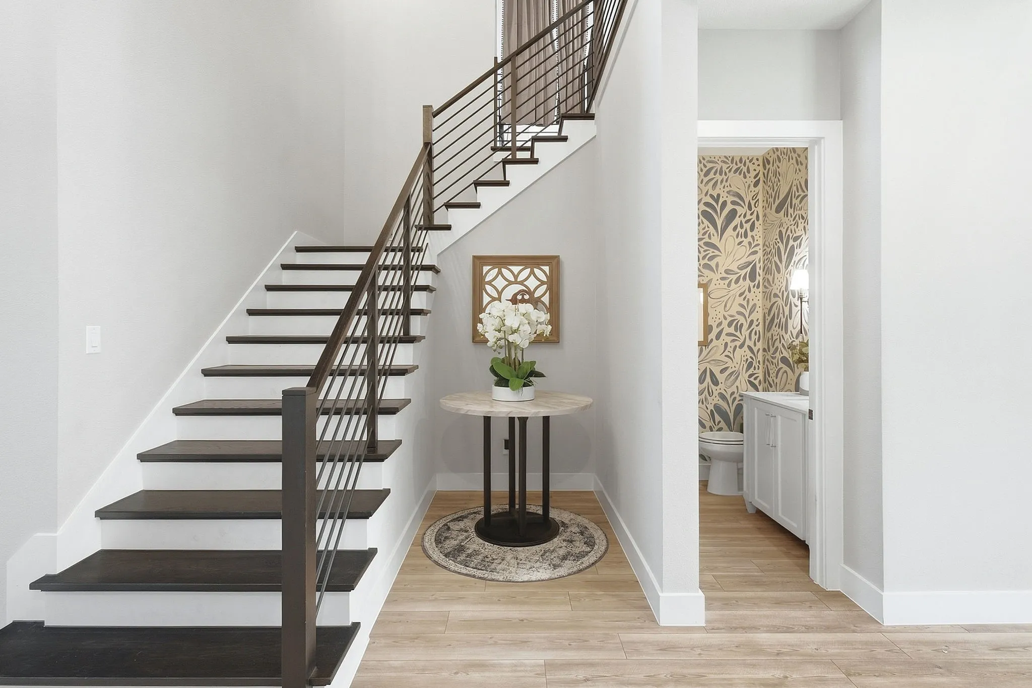 Hardwood flooring throughout the entry, staircase with dark wood treads and metal balusters, and a powder room featuring a pedestal sink and decorative wallpaper