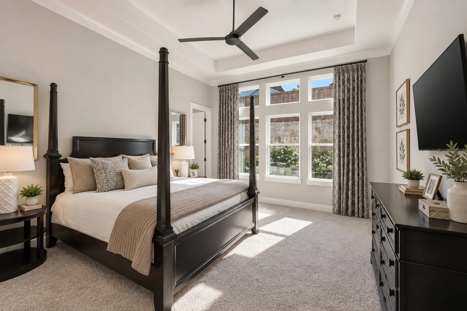 Recessed tray ceiling with crown molding, ceiling fan, large multi-pane windows, wall-mounted television, and light-toned carpet