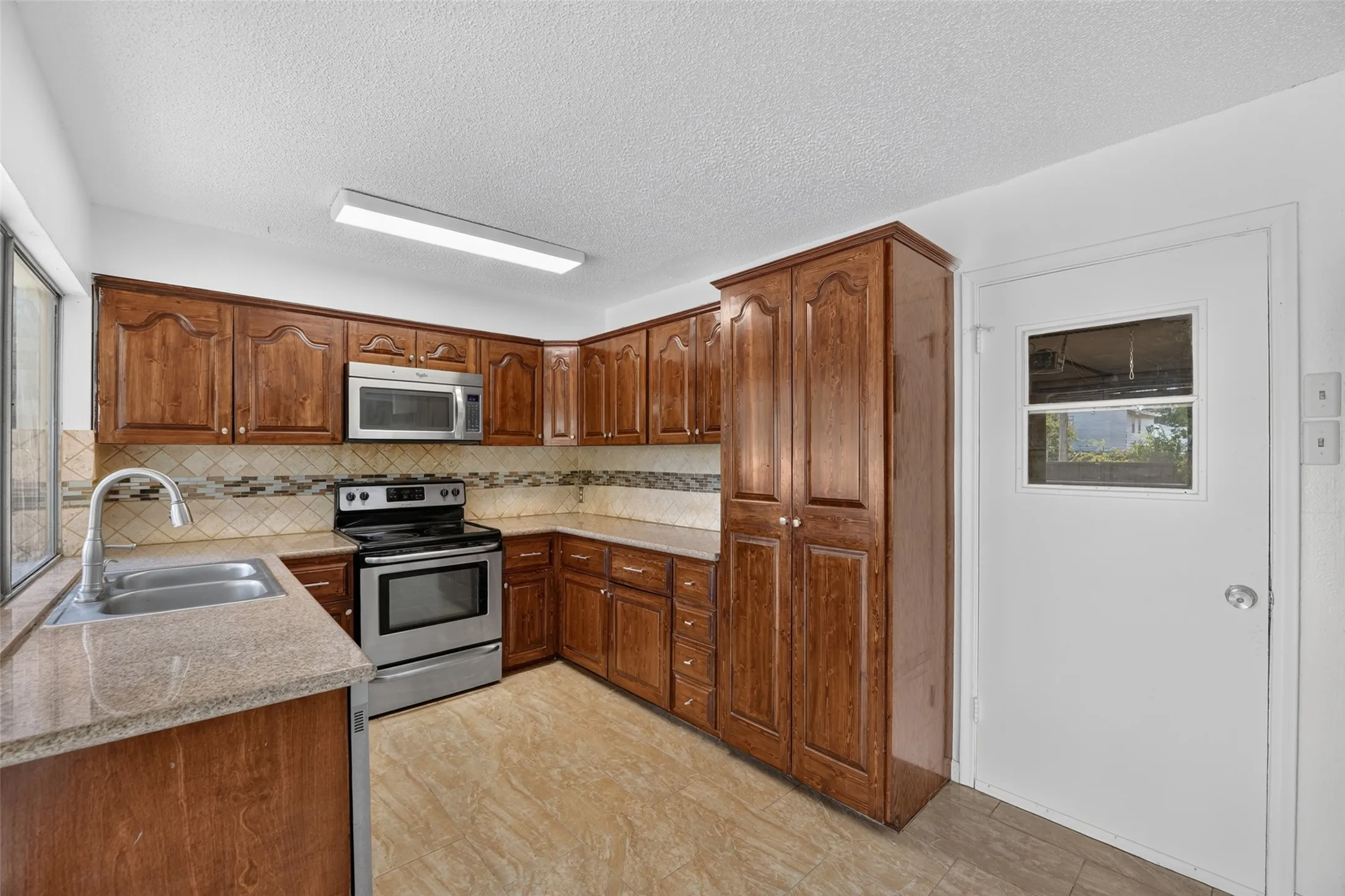 Kitchen featuring wood cabinetry, stainless steel appliances, patterned tile backsplash, double basin sink, and light-toned flooring