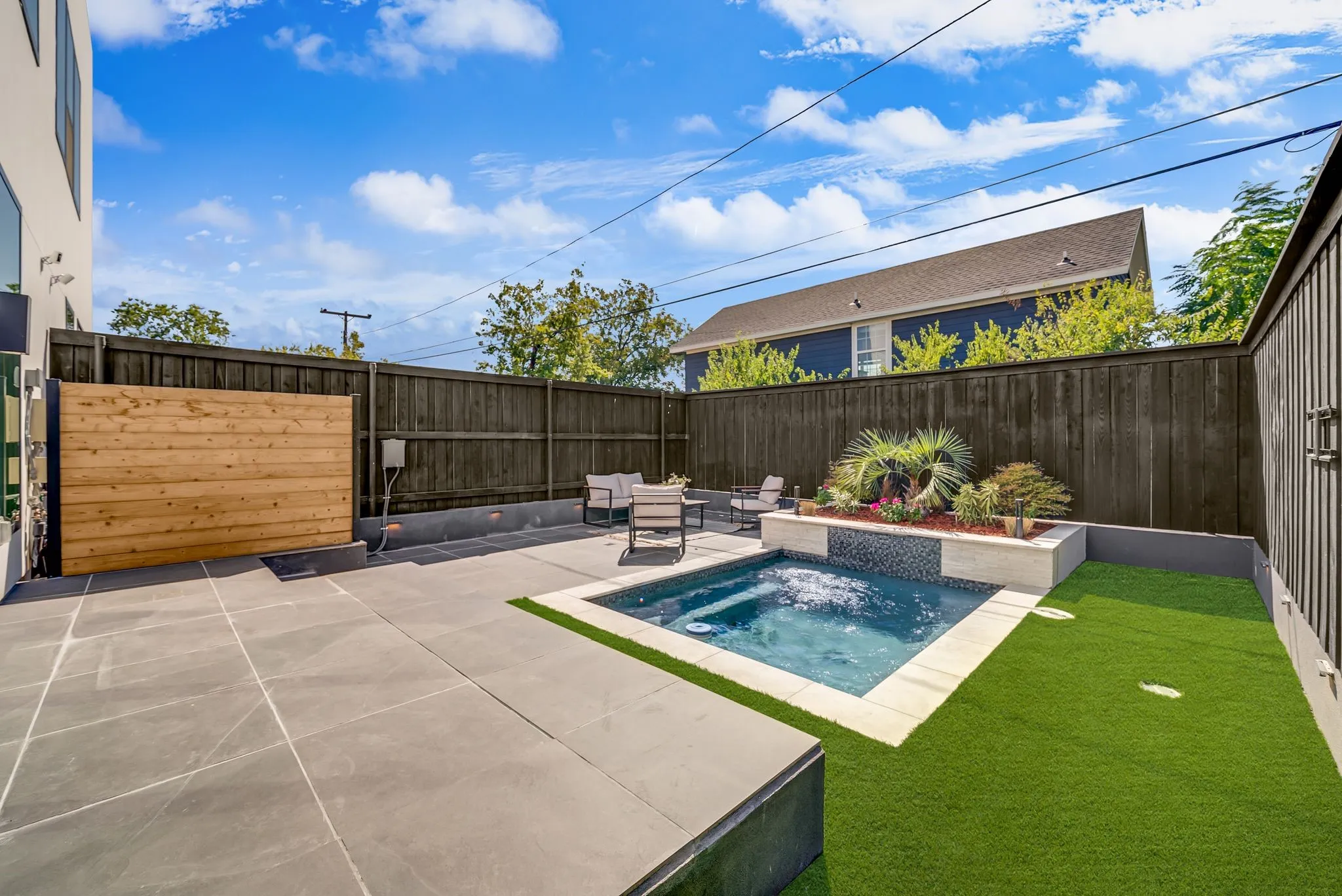 Private backyard featuring a built-in spa, dark privacy fencing, large format patio pavers, a planted raised garden bed, and artificial turf